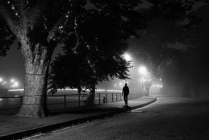 Une silhouette solitaire marche la nuit le long d’un chemin brumeux et faiblement éclairé au bord d’une rivière, aux prises avec des pensées de dépendance. Les lampadaires illuminent la scène brumeuse, projetant des ombres sur le trottoir. Des arbres ornés de guirlandes lumineuses bordent le sentier, contribuant à l’atmosphère à la fois étrange et sereine.