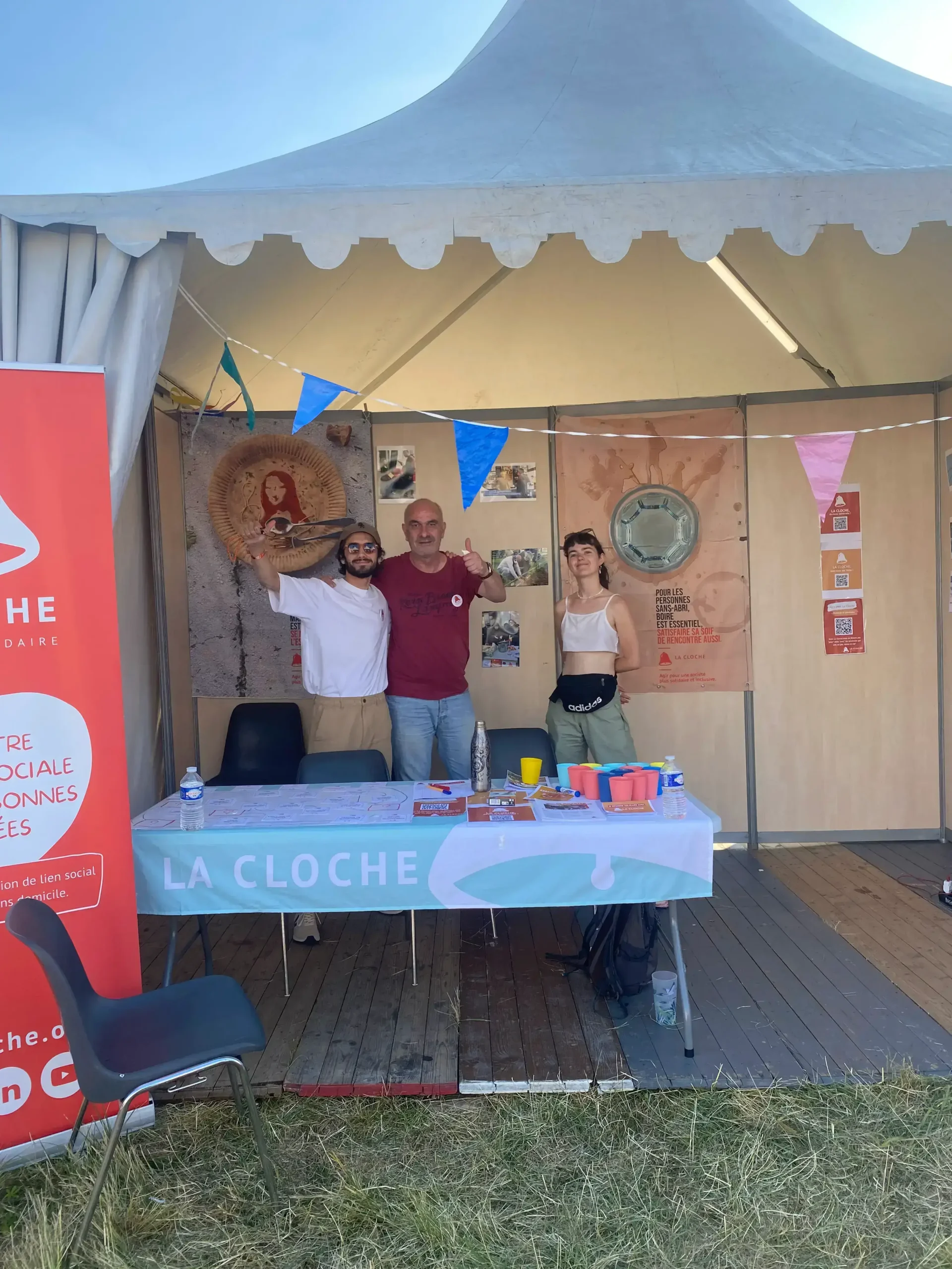 Trois personnes devant le stand de l'association La Cloche au festival Solidays 2023, présentant des actions de solidarité et de création de lien social avec les personnes sans-abri.