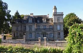 Image d'un manoir ancien en pierre, entouré de verdure et d'un jardin, avec des arbres en arrière-plan et un ciel bleu.